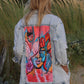 Light blue denim jacket with a rough finish and artwork of a woman's face on the back in pink, orange, and blue hues, featuring roses and a peace sign