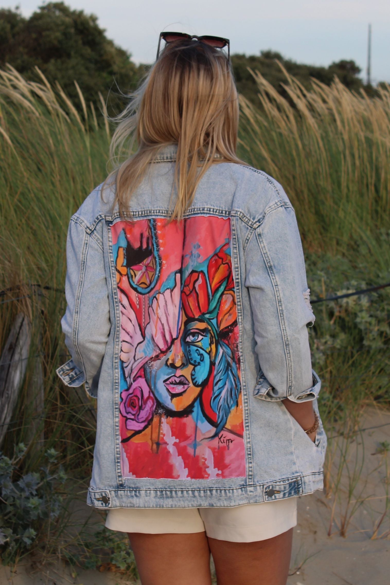 Light blue denim jacket with a rough finish and artwork of a woman's face on the back in pink, orange, and blue hues, featuring roses and a peace sign