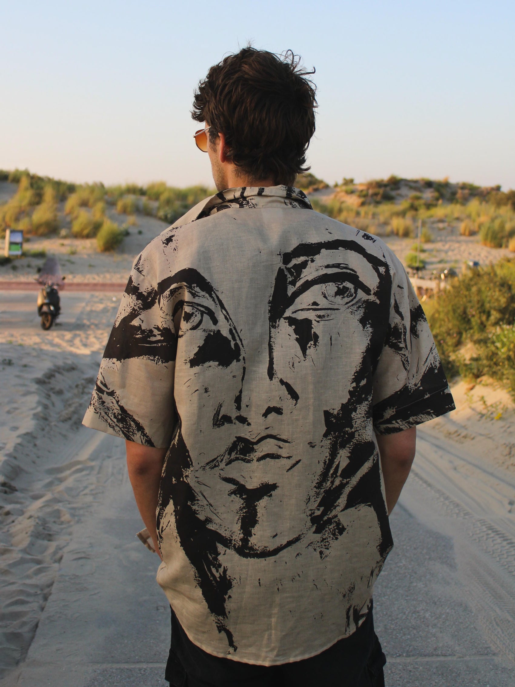 A man viewed from behind wearing a short-sleeve button-up shirt with a large black graphic face print on a light beige background, set against a sandy beach background.