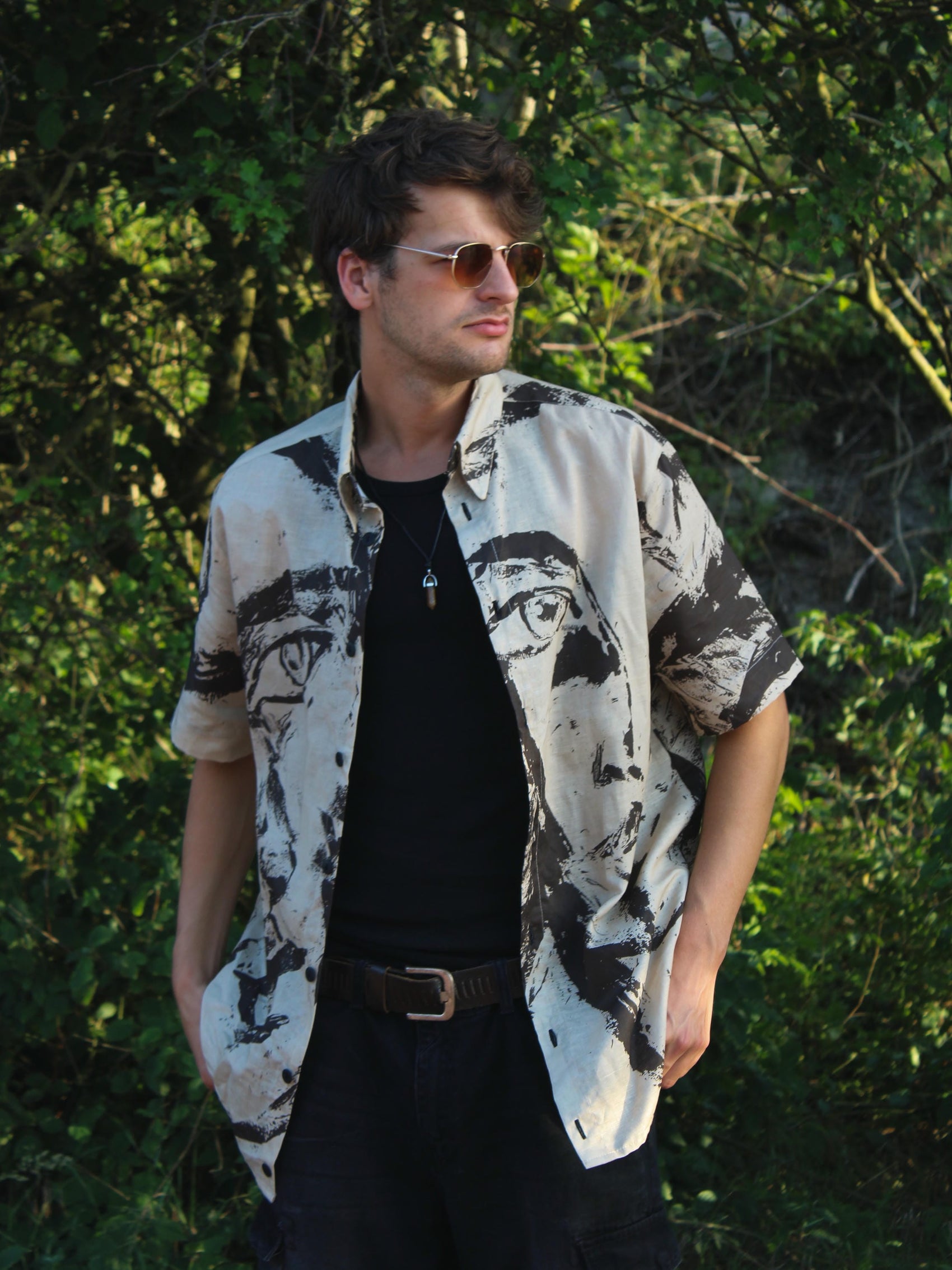 A man wearing a short-sleeve button-up shirt with a black and brown portrait print, styled over a black t-shirt and dark pants, standing outdoors against a background of green foliage.