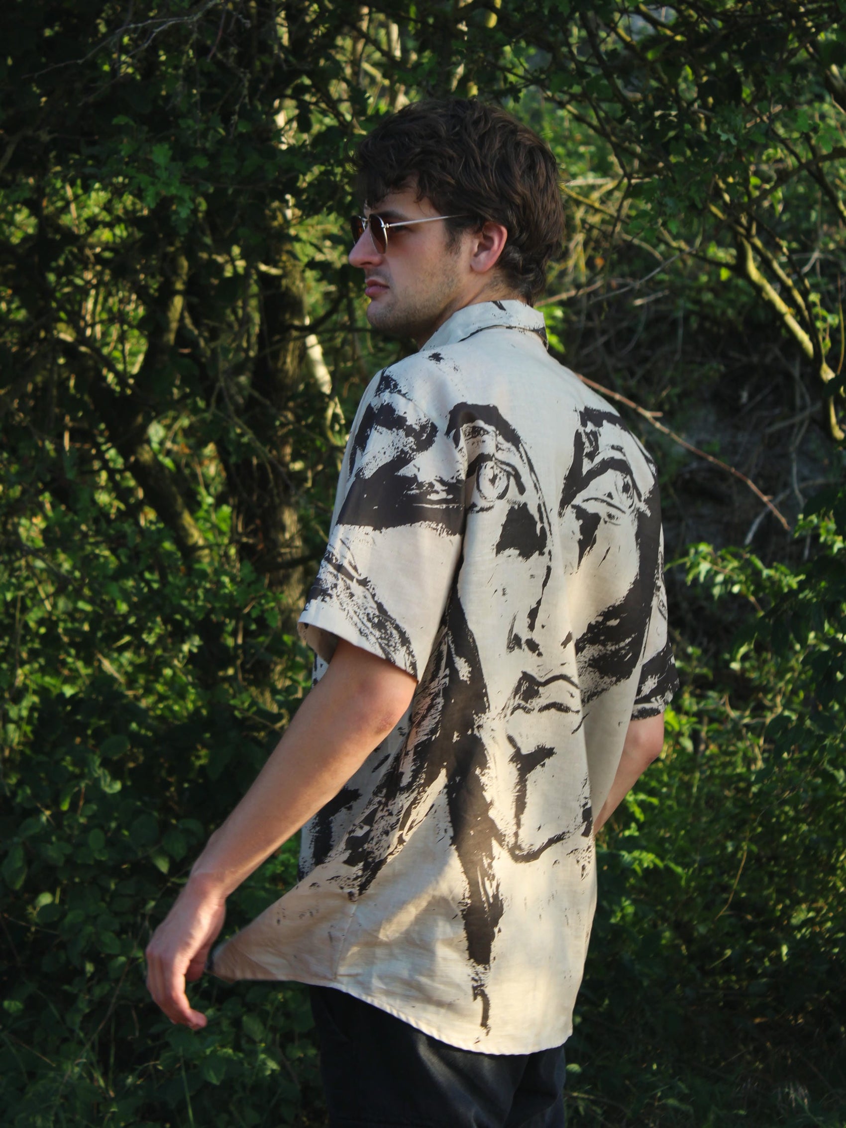 A man wearing a short-sleeve button-up shirt with a large black ink-style portrait print on an off-white background, standing outdoors in front of dense green foliage.