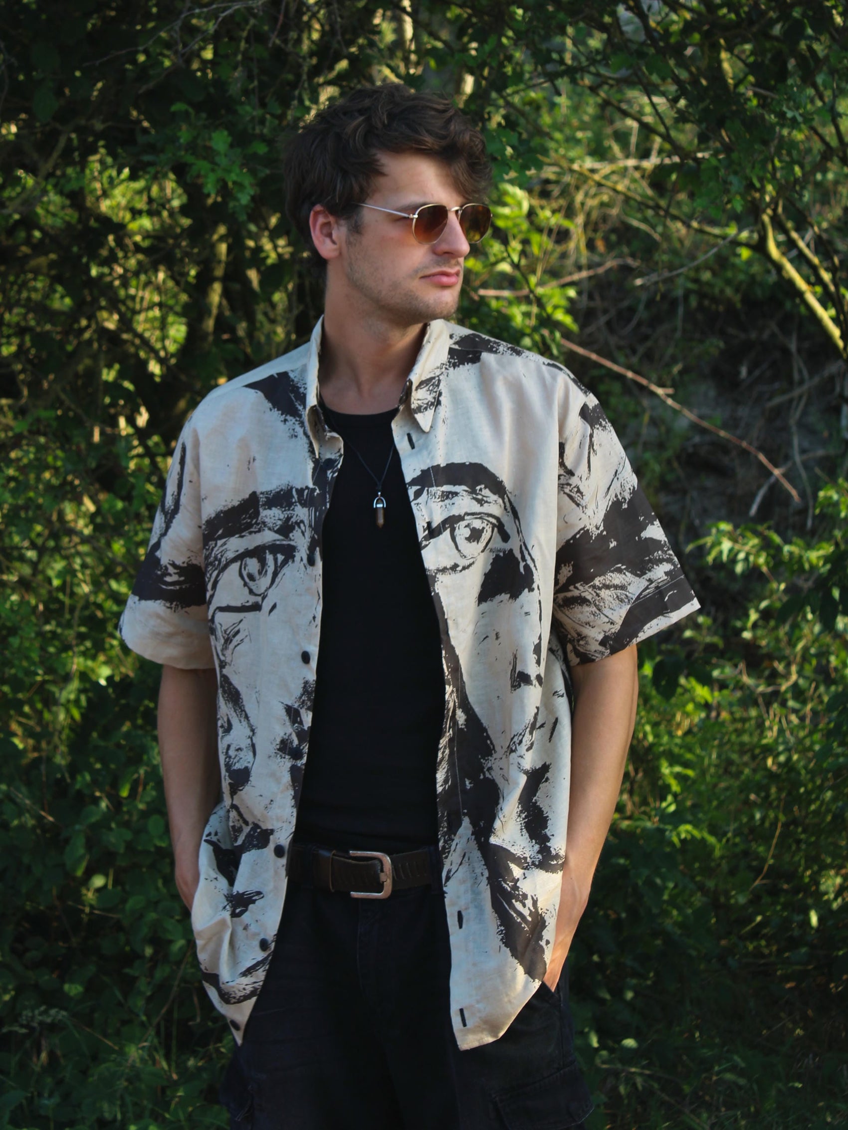 A man wearing a black and brown abstract art print short-sleeve button-up shirt over a black t-shirt, standing outdoors in front of a green forest background.